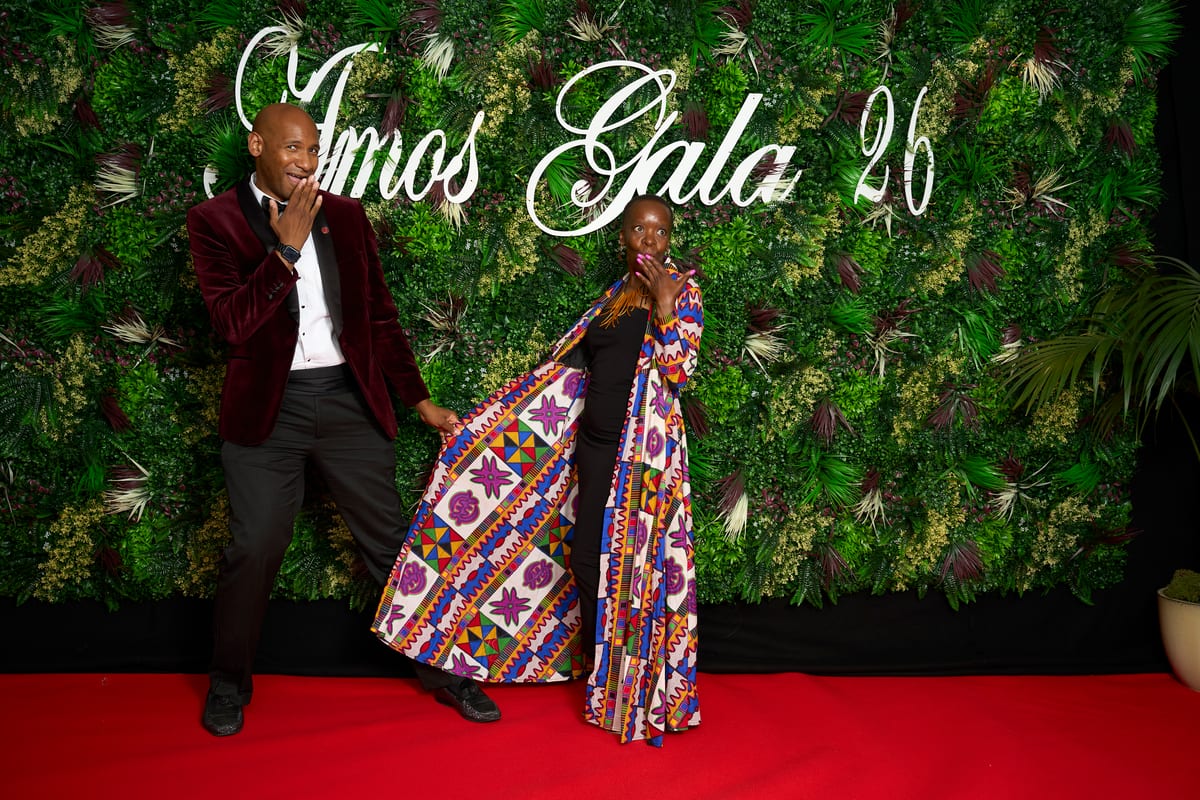 Two guests strike a playful pose on the red carpet at the Amos Gala, one in a burgundy velvet jacket and the other in a vivid patterned coat, against a wall of greenery