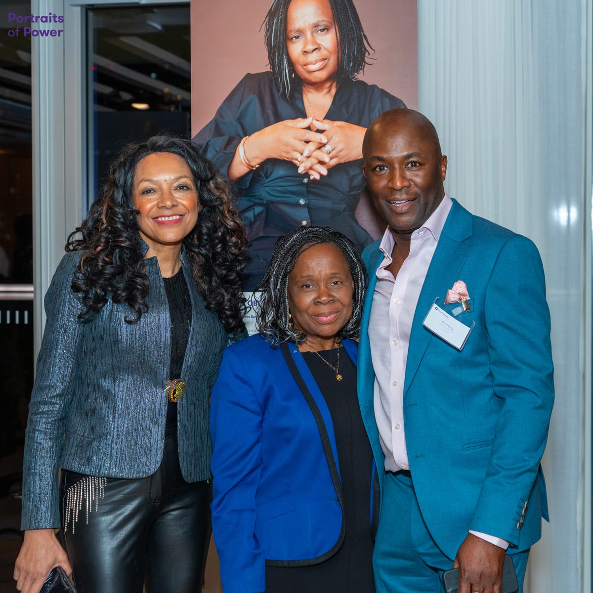 Three guests smile together at an evening event, in front of a large printed portrait on display