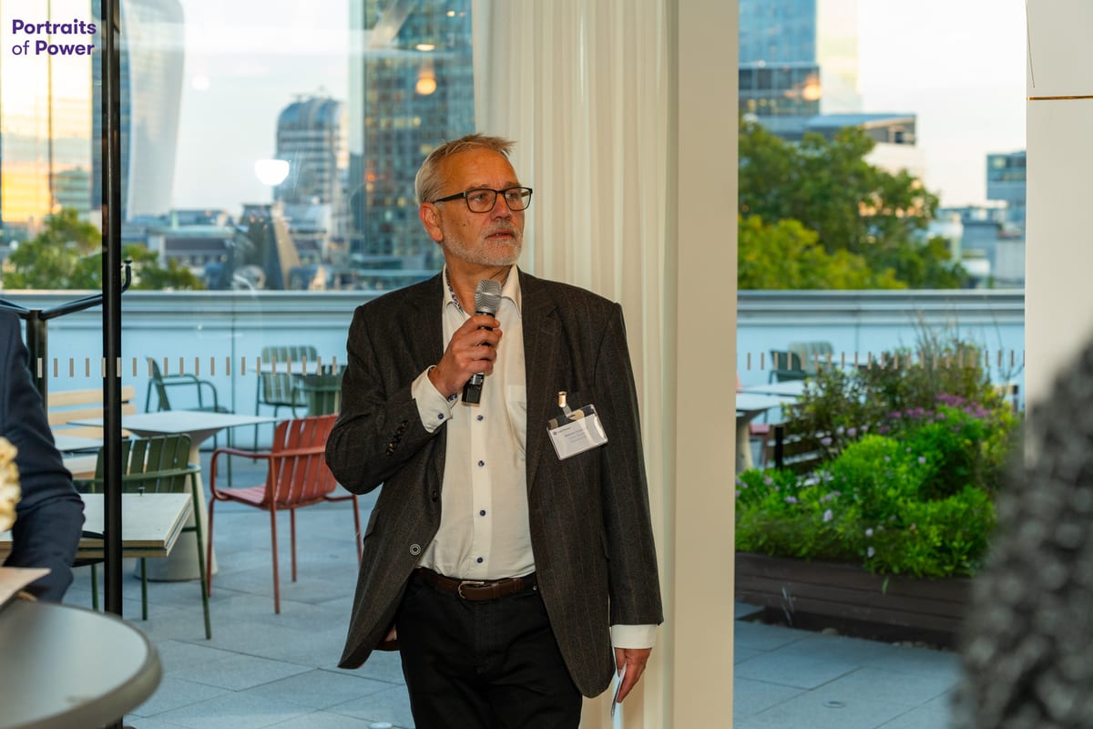 A speaker holding a microphone addresses guests at an evening event, with the London skyline visible through the windows behind him