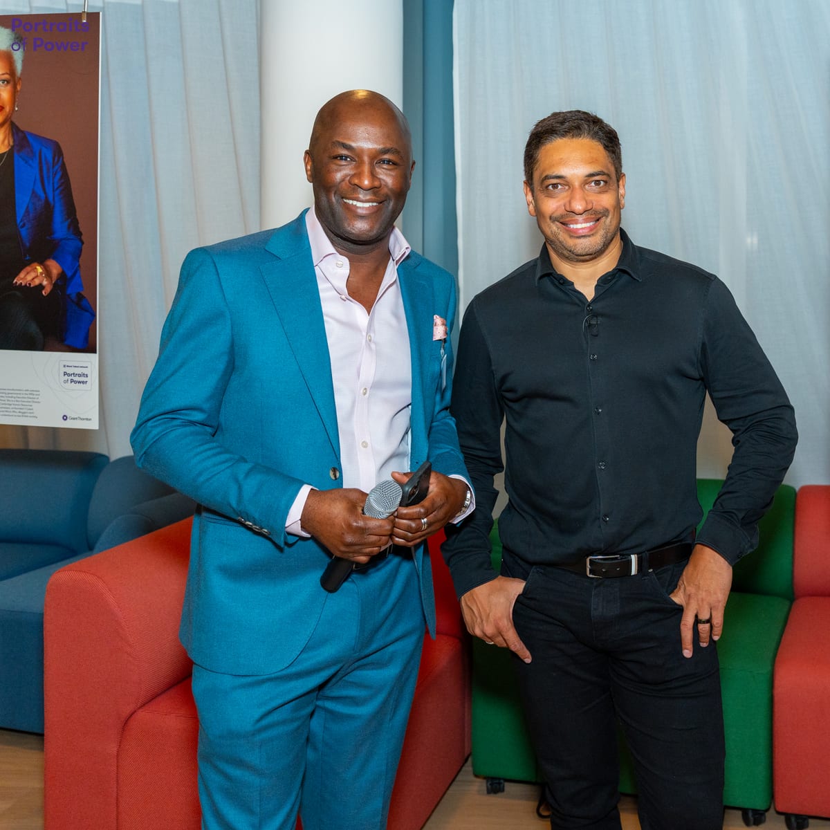 Two men smile for the camera at a corporate networking event, one in a bright teal suit holding a microphone