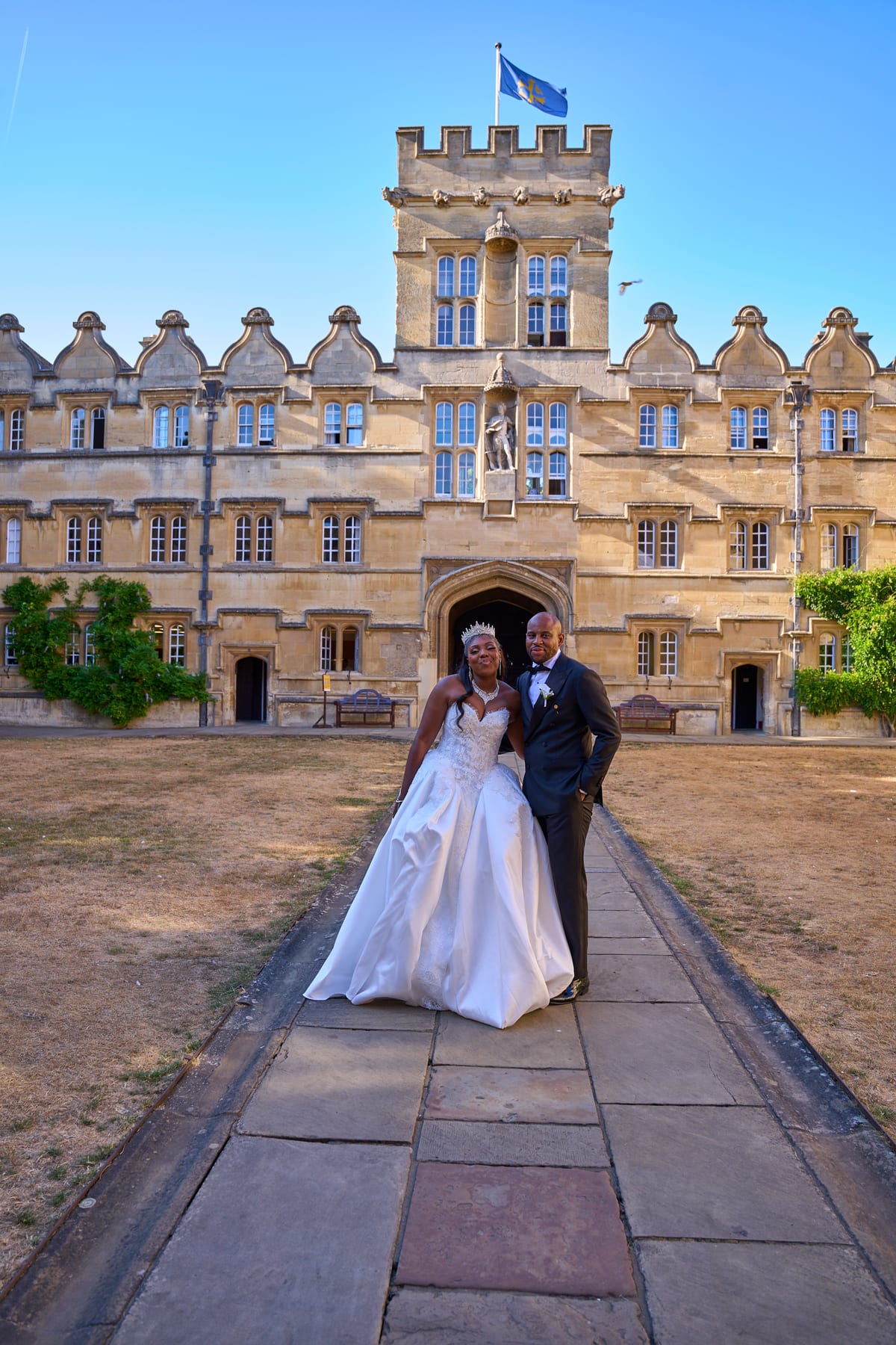 A bride in a tiara and white satin gown stands with her groom in a black tuxedo on a stone path in front of a historic college building, under a clear blue sky