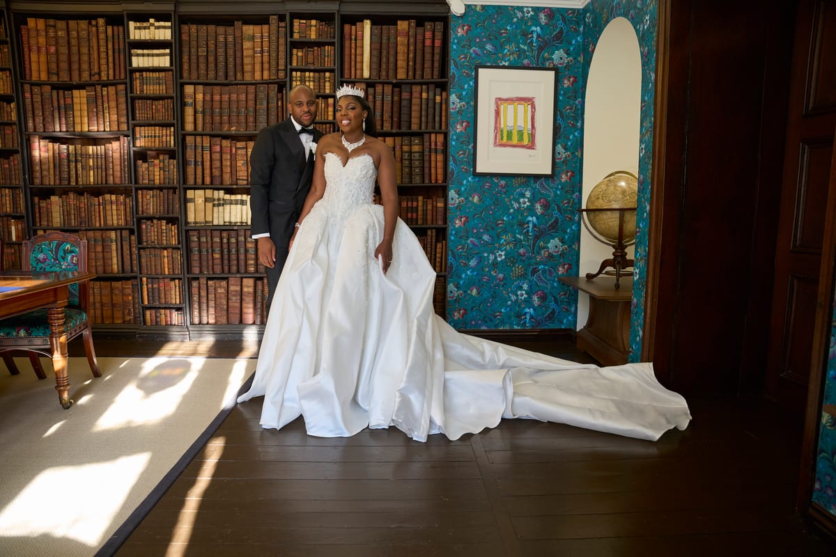 A bride and groom stand in a wood-panelled library lined with old books, her long satin train spread across the floorboards in the afternoon light