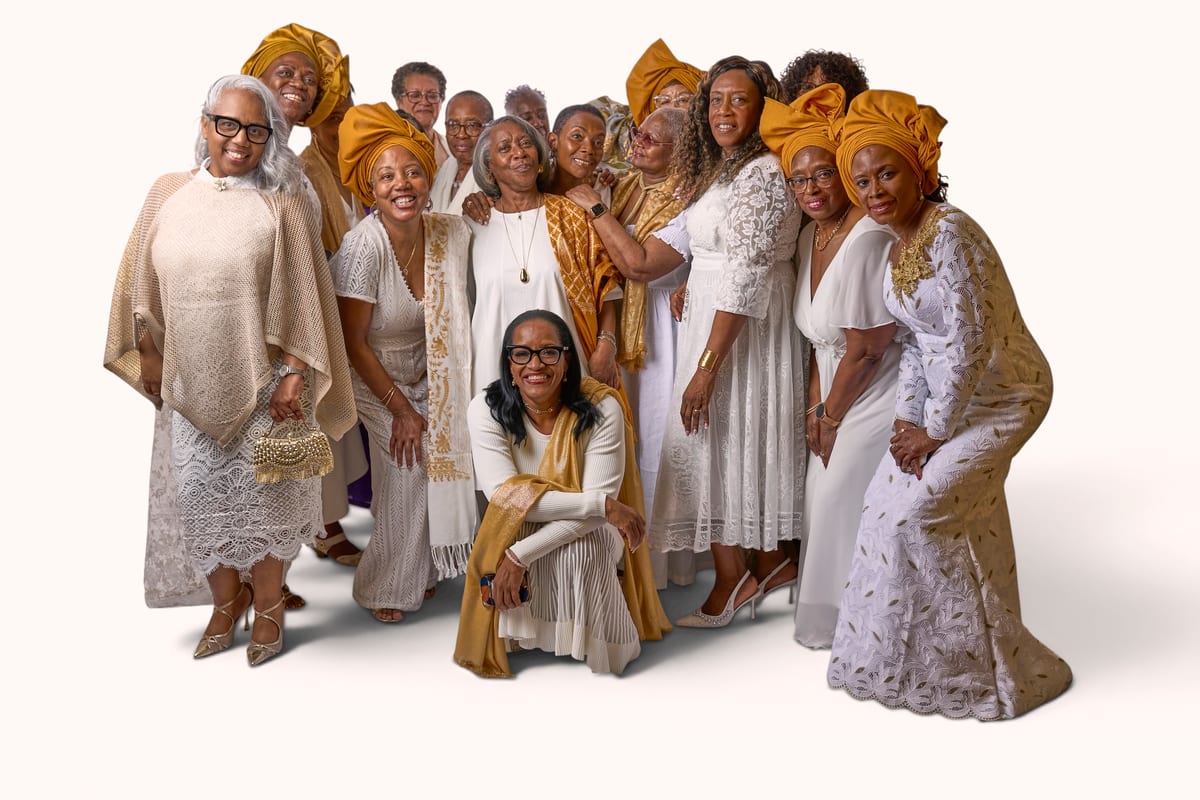 A group of women in white lace and gold, several wearing gold head wraps, pose together for a studio portrait
