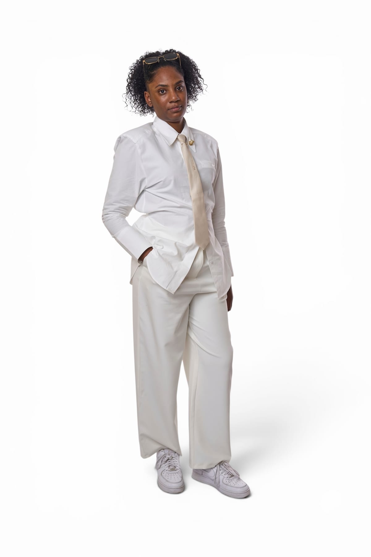 A full-length studio portrait of a woman in a white shirt, cream tie and wide white trousers, against a white backdrop