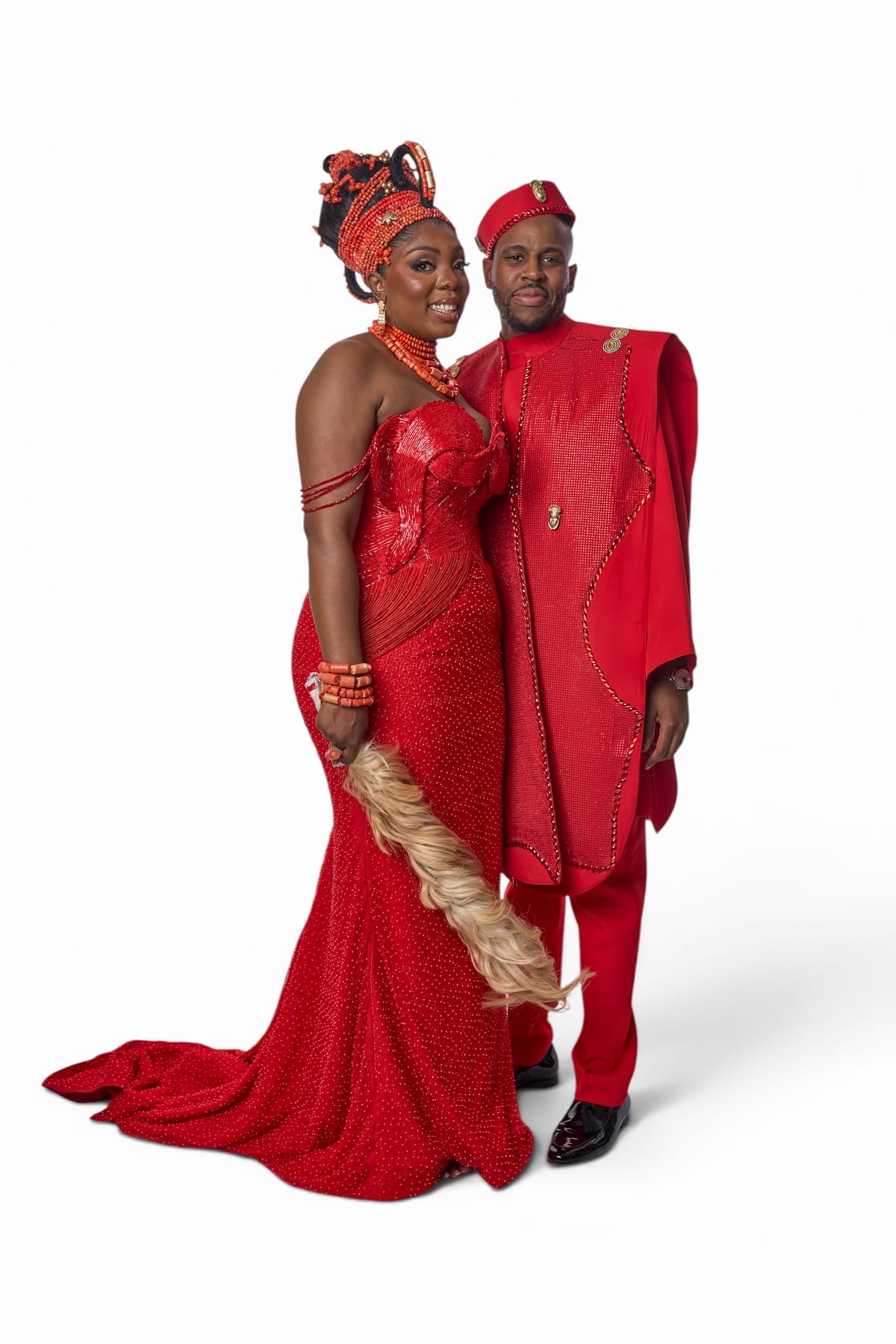 A couple in matching red traditional attire pose for a studio portrait, she in a beaded gown with coral bead jewellery and headdress