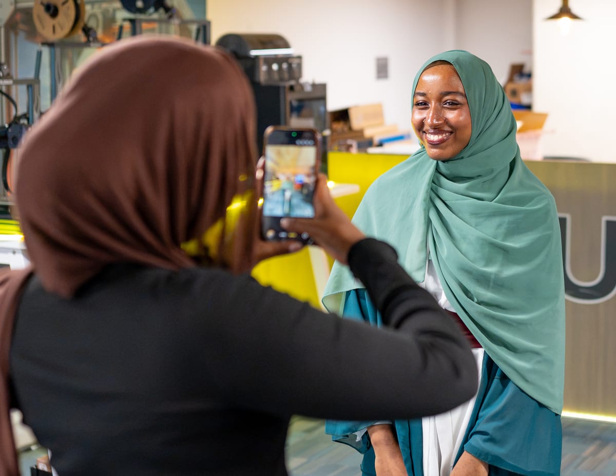 A smiling young woman in a green headscarf is photographed by a friend on a phone